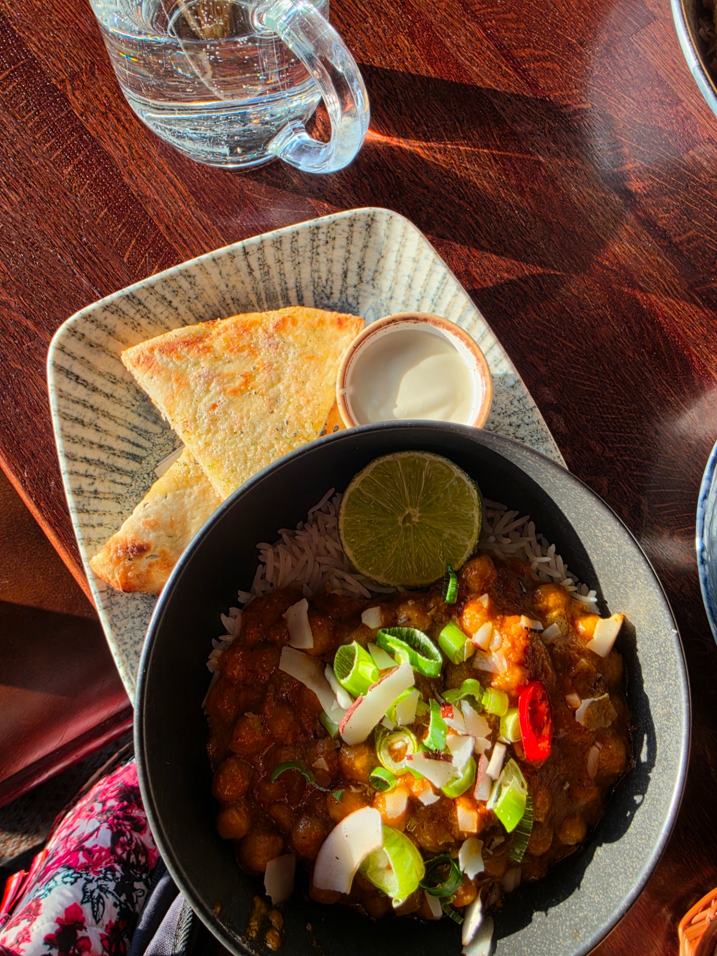 Kulhollinen curryä, riisiä ja limeviipale, rieskamallista leipää lautasella sekä lasillinen vettä puupöydällä.